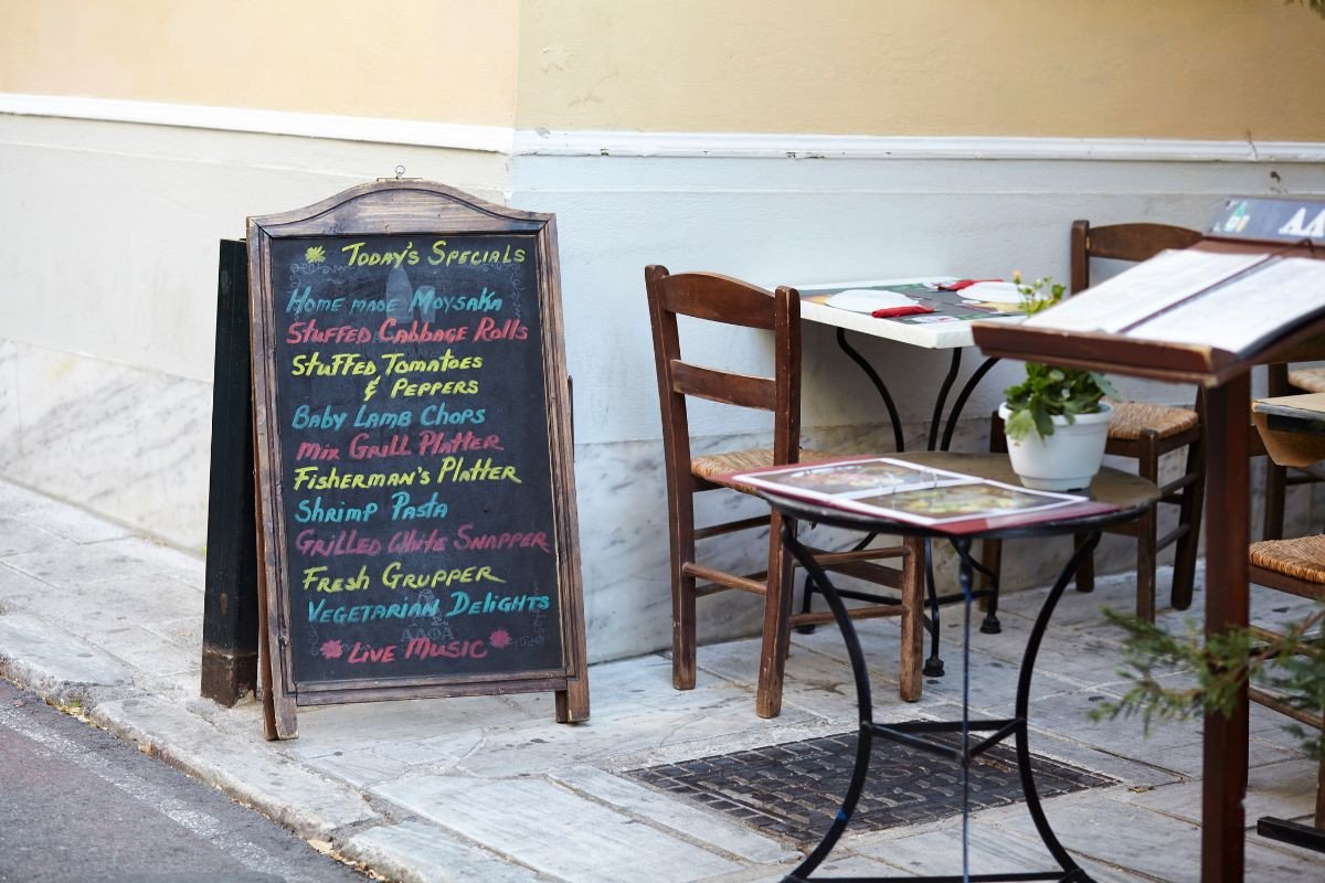A chalkboard outside a restaurant displays the day's specials in colorful chalk, including dishes like homemade moussaka, stuffed cabbage rolls, baby lamb chops, and grilled snapper. The board is positioned on a sidewalk next to a small table and chairs, creating an inviting atmosphere. The table has a menu and a small potted plant, while the restaurant’s wall is painted a soft yellow. The scene suggests a cozy and charming dining experience.